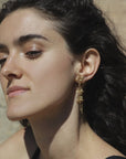 Close-up of a woman wearing a gold-plated floral dangling earring, handmade by Izaskun Zabala, with a blurred natural background.