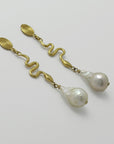 Pair of golden spiral earrings with baroque pearl drops, handmade by Izaskun Zabala, on a light gray background.