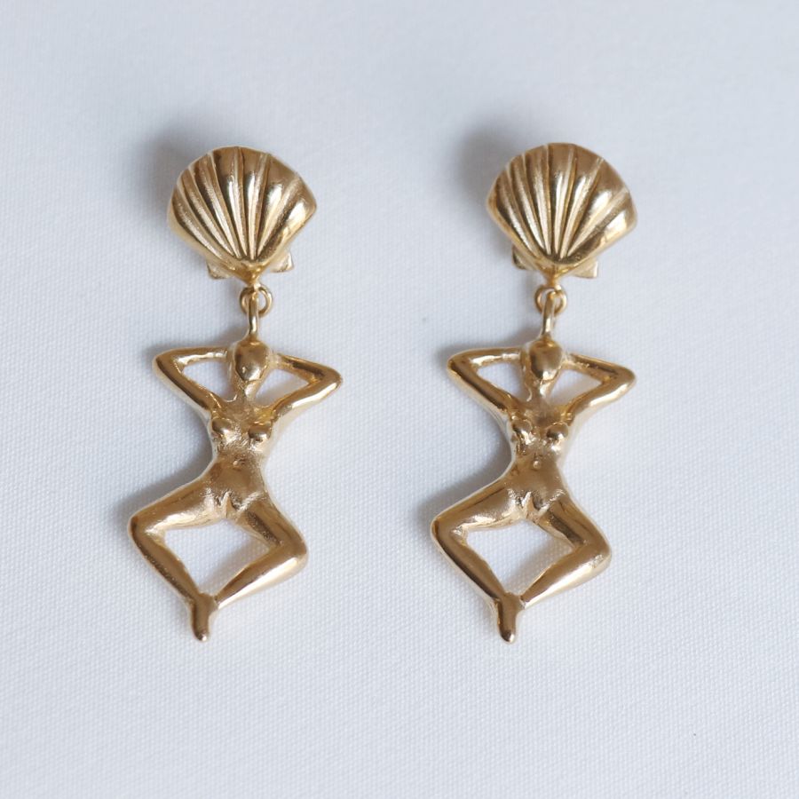 Image is a pair of earrings of a woman's figure dangling from a shell stud, handmade by Izaskun Zabala.