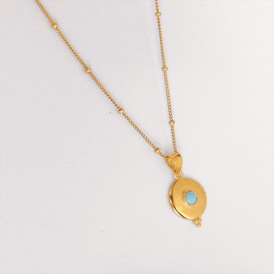 Image is a golden disc pendant with a turquoise cabochon center stone and a dainty chain, handmade by Izaskun Zabala.