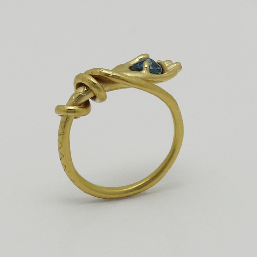 Image is a 24K gold-plated snake like band with a hand holding a topaz ring, handmade by Izaskun Zabala.