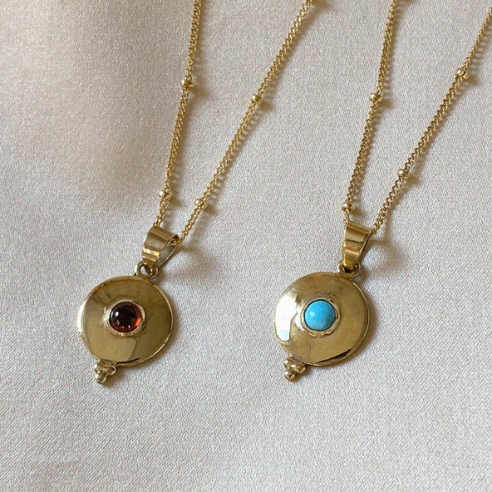 Image is a round brass pendant with a center stone cabuchon and a gold filled chain, handmade by Izaskun Zabala.