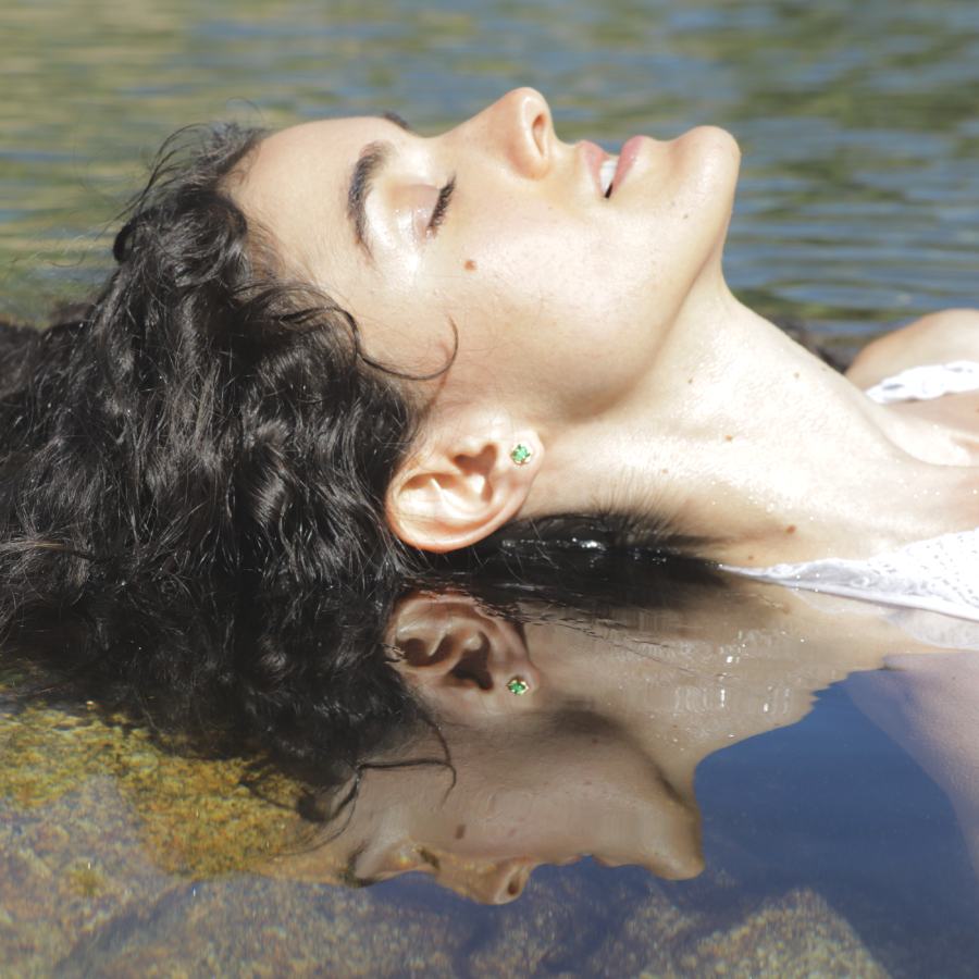 Model lying on water with reflection, wearing an emerald hexagon stud earring, handmade by Izaskun Zabala.