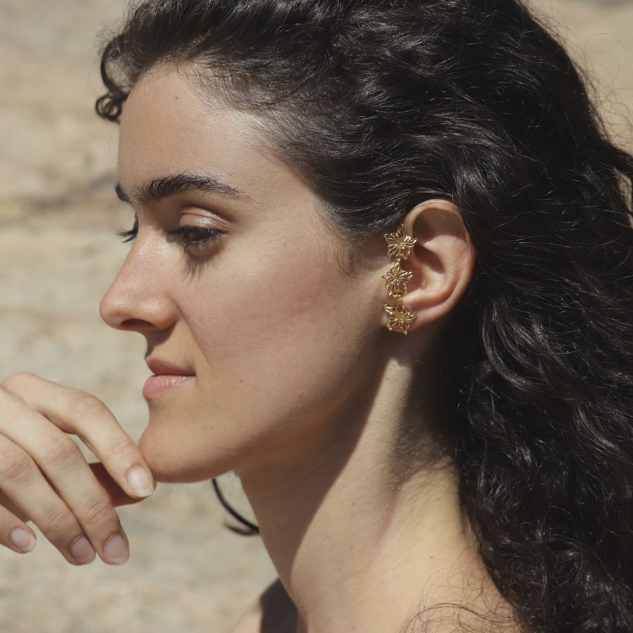 Close-up of a woman wearing a golden floral climber earring, handmade by Izaskun Zabala, with a blurred natural background.