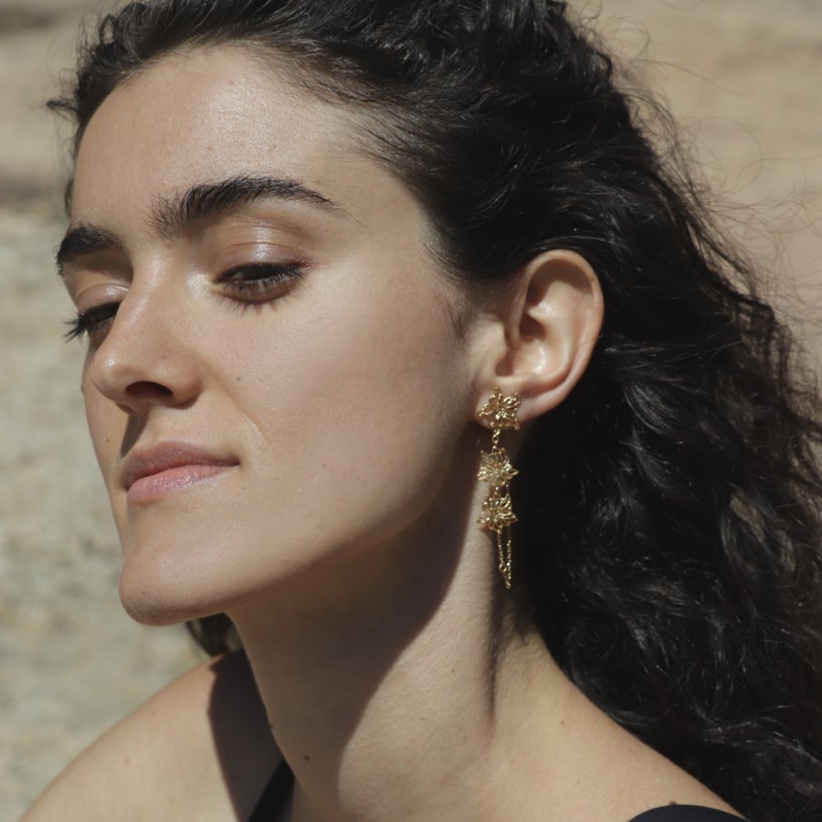 Close-up of a woman wearing a gold-plated floral dangling earring, handmade by Izaskun Zabala, with a blurred natural background.