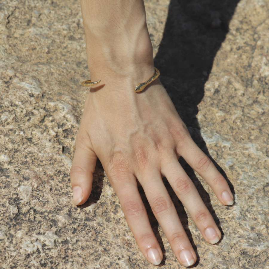 Hand with a golden snake-shaped bangle handmade by Izaskun Zabala, on a textured stone surface.