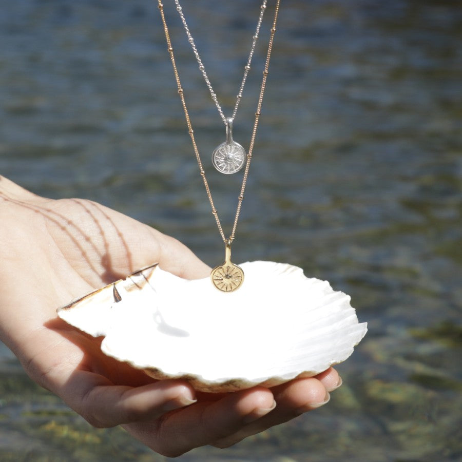 Hand holding a shell with carved sun medallion layered necklaces, handmade by Izaskun Zabala, against a water background.