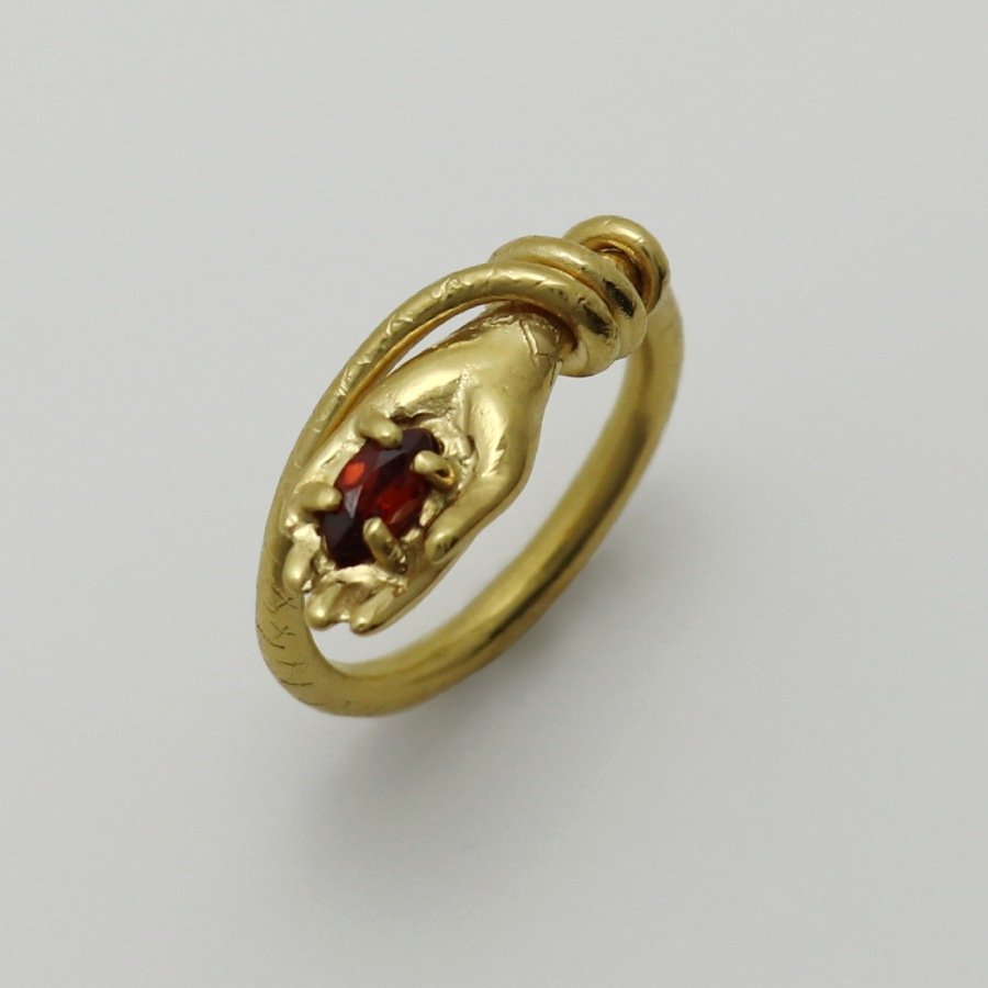 Image is a 24K gold-plated snake like band with a hand holding a garnet ring, handmade by Izaskun Zabala.