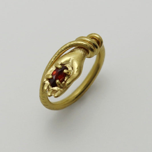 Image is a 24K gold-plated snake like band with a hand holding a garnet ring, handmade by Izaskun Zabala.