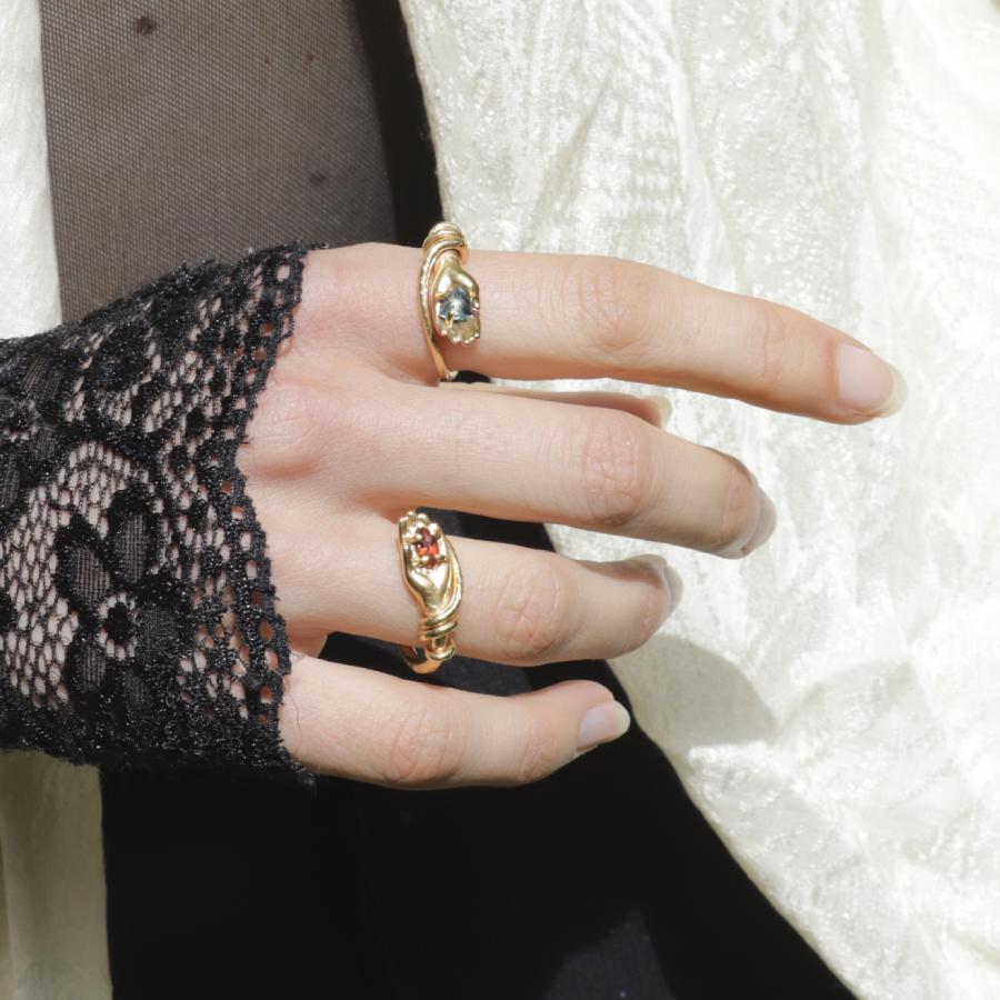 Image is a pair of 24K gold-plated snake like band rings with a hand holding a gemstone, handmade by Izaskun Zabala.
