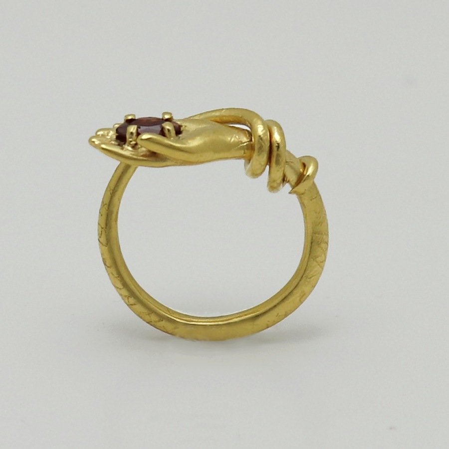 Image is a 24K gold-plated snake like band with a hand holding a garnet ring, handmade by Izaskun Zabala.