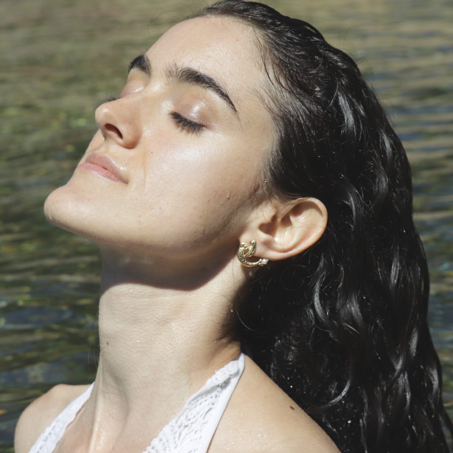 Woman with wet hair wearing a golden mermaid stud earring against a water background, handmade by Izaskun Zabala.