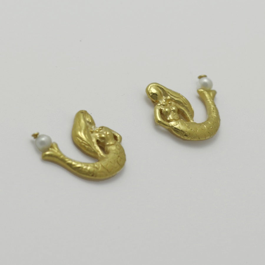 Golden mermaid-shaped earrings with freshwater pearls on a light gray background, handamade by Izaskun Zabala.
