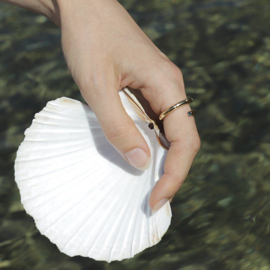 Gold plated opened band with blue gemstone at the end, handmade by Izaskun Zabala, on a hand holding a white shell.