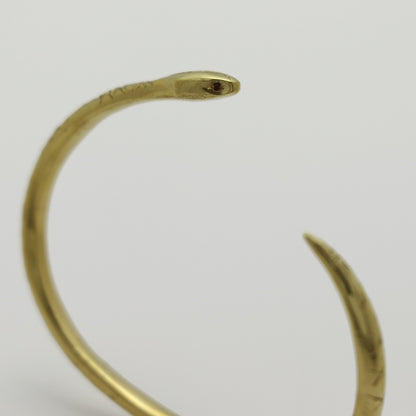 Golden snake-shaped bangle with garnet eyes handmade by Izaskun Zabala, on a light gray background.