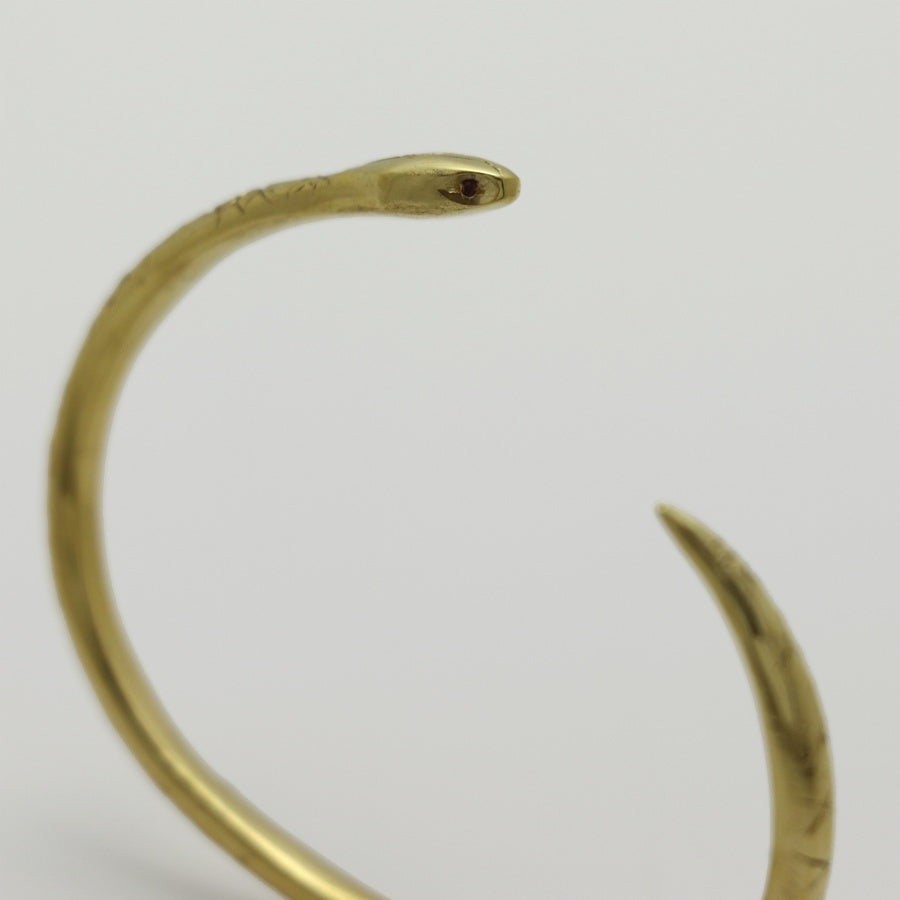 Golden snake-shaped bangle with garnet eyes handmade by Izaskun Zabala, on a light gray background.