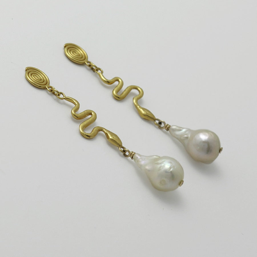 Pair of golden spiral earrings with baroque pearl drops, handmade by Izaskun Zabala, on a light gray background.