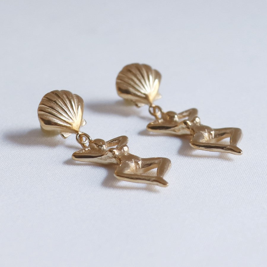 Image is a pair of earrings of a woman's figure dangling from a shell stud, handmade by Izaskun Zabala.