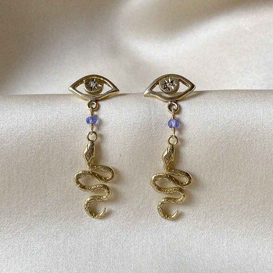Image is a pair of eye studs with a dangling serpent from a tanzanite stone, handmade in brass by Izaskun Zabala.