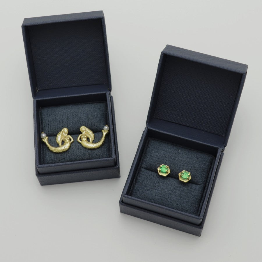 Two pairs of golden unique earrings in dark blue Izaskun Zabala branded boxes on a light gray background.