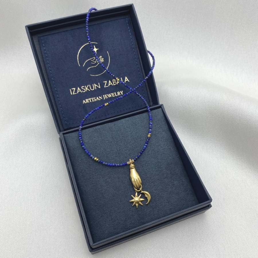 Image is a lapis lazuli beaded chain necklace with a carved hand, sun and moon gold-plated pendant, displayed in the brand&#39;s packaging.
