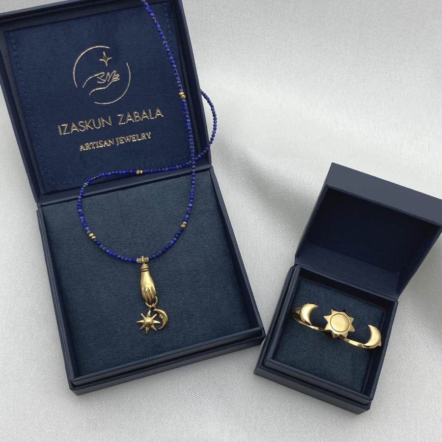 Image is a lapis lazuli beaded necklace and a double finger moon and sun golden ring, displayed in the brand&#39;s packaging.