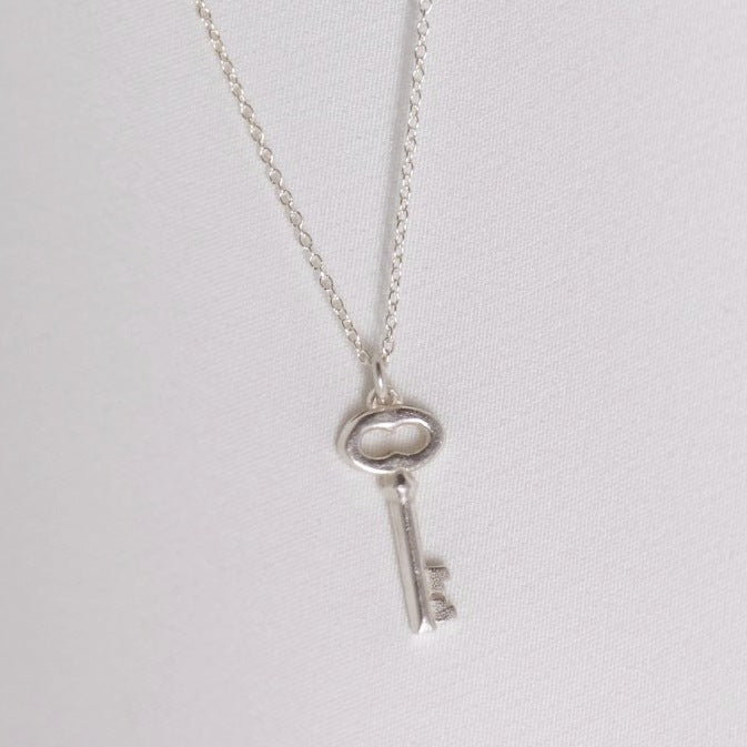 Image is a silver key charm pendant delicate necklace, handmade by Izaskun Zabala.