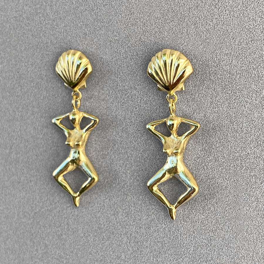 Image is a pair of earrings of a woman's figure dangling from a shell stud, handmade by Izaskun Zabala.