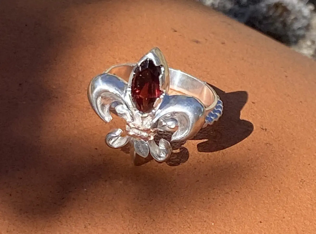 A custom sterling silver fleur-de-lis ring featuring a marquise-cut garnet gemstone, displayed on a textured terracotta surface with direct sunlight casting a strong shadow.