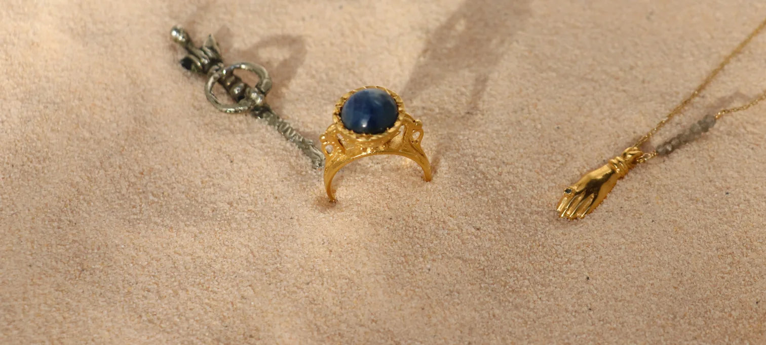 Gold-plated cabochon statement ring and palmistry pendant on sand, artisan-made by Izaskun Zabala.
