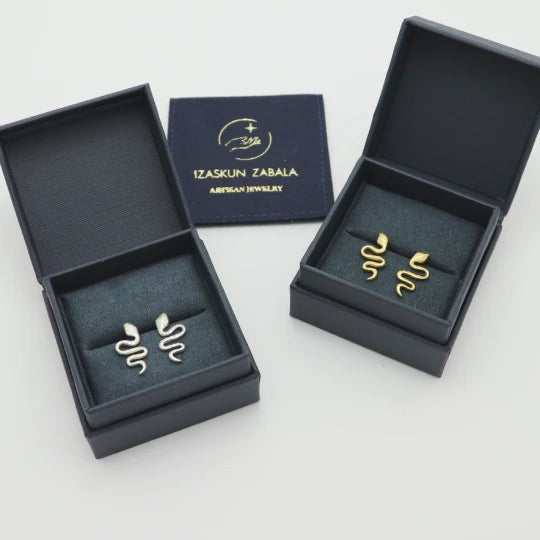 Gold-plated snake-shaped earrings in open dark blue boxes with 'Izaskun Zabala' branding.