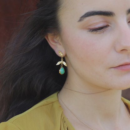 Close-up of a woman wearing gold-plated leaf earrings with green teardrop stone.